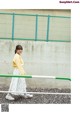 A woman in a yellow sweater and white skirt standing next to a fence.