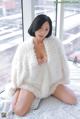 A woman sitting on a bed in a white fur coat.